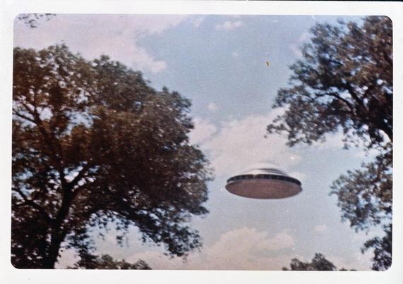 Historic WikiCommons color photographic image from 1964 of a "Flying saucer from the USA National Archives and Records Administration". The day is sunny with blue skies & white clouds. We are looking up between 2 large trees. Typical silver flying saucer shape with a row of small dark windows around its middle. 

Dated 1964 Jun 04, Author Unknown. 
https://commons.wikimedia.org/wiki/File:Flying_Saucer_-_NARA_NAID_303938034.tif 

www.KronoMoon.org
#KronoMoon