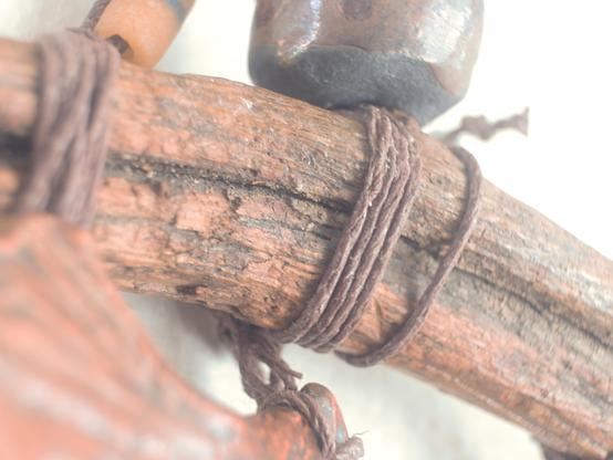 A close-up view of the same wall hanging focuses on the coarse, reddish-brown branch at the top, tightly wrapped with multiple strands of brown string. Small, smooth beads of varying shapes and shades rest beside the twine, while the detailed texture of the wood’s grain and the fibrous strands of the cord fill the frame. The background is softly blurred, emphasizing the tactile details.