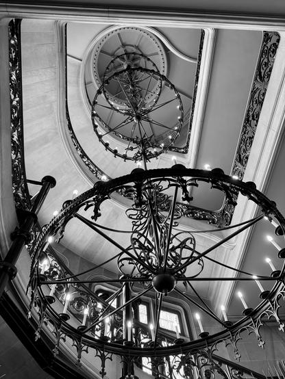 Black and white image looking up at three circular iron chandeliers with white candles suspend from the top of a staircase. The staircase also has ornate iron rails.