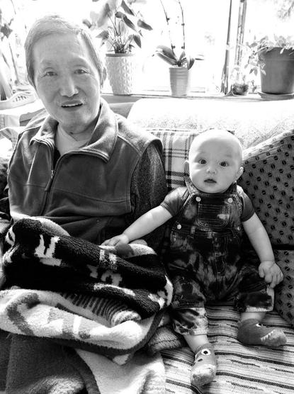 A grayscale image shows an older Asian man and a baby sitting together on a patterned couch in front of a window with potted plants. The man, wearing a vest over a sweater, smiles at the camera. The baby, wearing denim overalls and socks, looks up with a surprised expression, holding the man's hand. A folded blanket rests near the man.