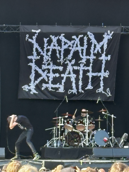 Kuvassa laulaja kumartuneena lavalla päätään suojellen ja taustalla rumpali soittamassa. Takana Napalm Death -yhtyeen logolakana.