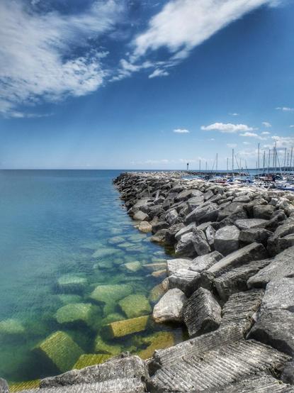 A large rock built breaker wall is seen stretching into the distance protecting a marina on Lake Huron. The blue sky with soft white clouds and the clear blue water is seen as well as the masts of several sailboats to the right of the photograph.