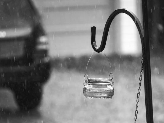 Black and white photograph capturing a glass mason jar suspended from a curved iron shepherd's hook during rainfall. Diagonal rain streaks streak across the frame while the jar hangs motionless with delicate wire handles. The background shows soft, blurred residential structures and natural elements, creating gentle layers of gray tones. Sharp focus emphasizes the jar's ribbed texture and metallic hardware against the atmospheric conditions.