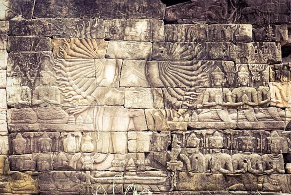 Photo shows an old carving of a figure with an enormous number of arms and hands, symbolising Lokeshvara, a Boddhisattva targeting helping humanity. Some details are very worn out, like the face, but most of the carvings are still very visible. Around the main figures there are rows of figures praying or bringing offerings to the Lokeshvara. There is a decorated lintel on top. Over the rows of prayers and behind the bodhisattva there are trees, temple and stupa towers.