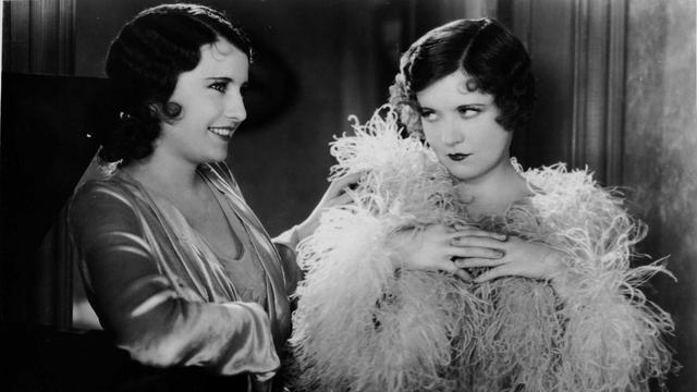 an image of two stylish young women, their hair wavy, one is wearing a shirt with extravagant feathering on it, the other grinning, looking to the right, theyre seen from their abdomens up