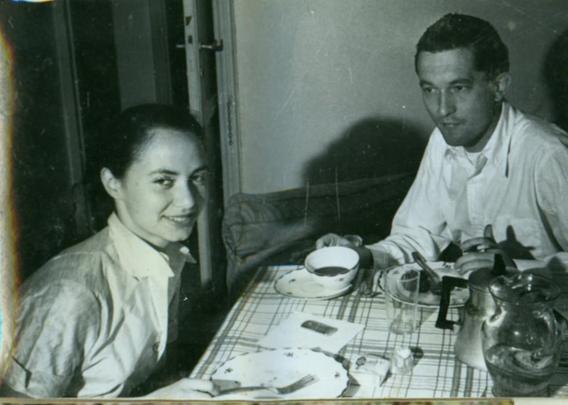 a painting of a woman and man sitting at a table, a cloth on it , there are some coffee pots and pitchers on the table, they are drinking coffee, she is looking at the viewer, he is looking at her, their shadows on the wall behind them