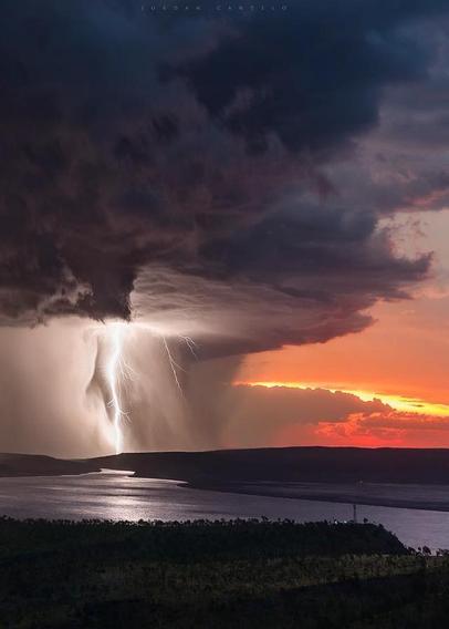 A photo of a lightning in Wyndham, WA by John Cantelo 

White lightning over smooth water, black storm cloud, orange-red sky.