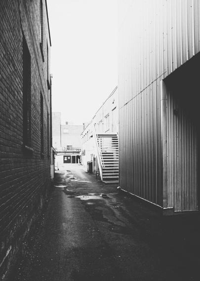 An alleyway behind shops and restaurants is seen in this high contrast black and white image. A large brick wall is seen on the left and a coragated steel wall on the right. Windows and a metal staircase can also be seen. The pure white sky is completely blown out.