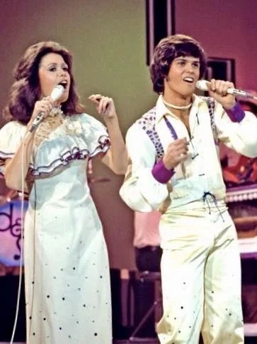 Donny and Marie Osmond sing together on their 1970s TV variety show. She looks a little bit country while he looks a little bit rock and roll.