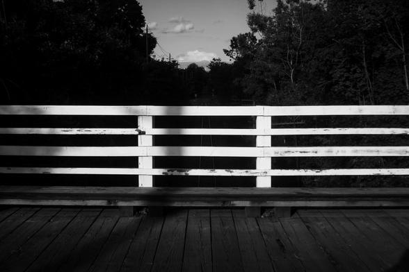 The white wood railing on an old road bridge glows in the early evening sunlight. The boards of the wooden bridge can be seen at the bottom of the image and the dark silhouette of trees and the sky containing a few clouds is seen in the background. This is a black and white image.