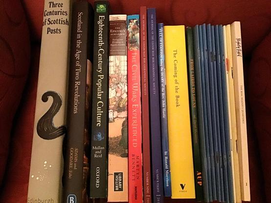 Lots of books lined up with their spines showing up to us, resting on a red chair. The books are varied, large, small, hardback, paperback, and many pamphlets. Subjects include things like "Three Centuries of Scottish Posts", "Scotland in the Age of Two Revolutions", and "The Coming of the Book".