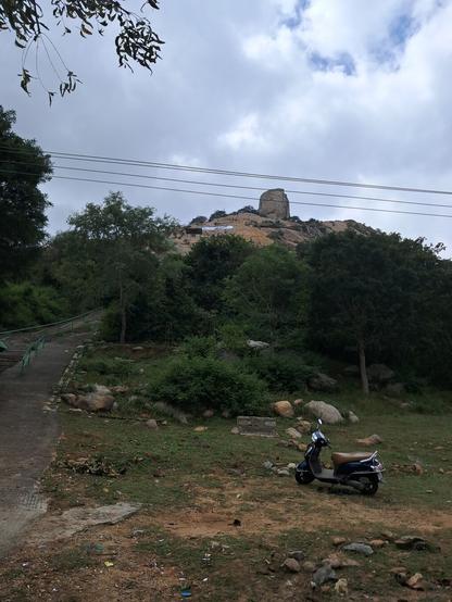 A large stone standing upright like a column on top of a hill center right of the image. Lower portions of the hill are green and covered with trees or grass. Upper portions of the hill are rocky and bare. To the left of the picture, a set of stairs with green railings leads up the hill. About halfway up the hill upon bare rock, there is a shed and above, a white rectangle is painted on the hill's rocky surface. Above the rectangle, the green railing goes horizontally from left to right and then goes up again, towards the column rock at the top. A plastic yellow water tank can be seen near the place where the railing goes up again.