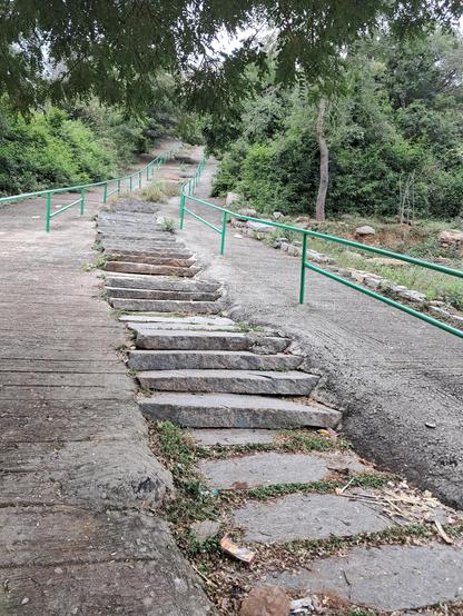 A rough staircase made of rocks slabs with concrete to sides, leading up a slope. Painted green railings are there on both sides of the staircase with posts embedded in concrete. Further up the slope, trees can be seen on both sides. Branches of a tree behind the camera overhang the top part of the image.