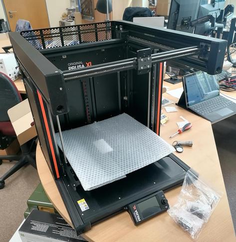 Foto eines 3D Filamentdruckers von der Marke Prusa, der sich im Aufbau befindet. Das Büro ringsum ist sehr unordentlich.