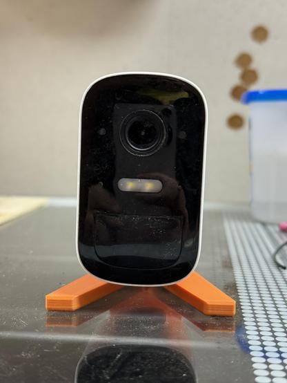 A white Eufy security camera on an orange 3D-printed stand, shown from the front indoors. The camera lens and sensor are clearly visible on the glossy black front panel, with a reflection of the photographer faintly visible.
