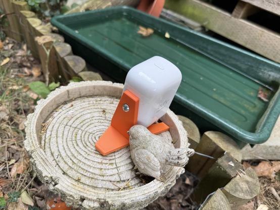 A small white Eufy security camera is mounted on an orange 3D-printed stand, placed on top of a wooden garden ornament shaped like a tree stump with a small bird figure attached. In the background, there is a green plastic container and wooden fencing with plants and leaves around.