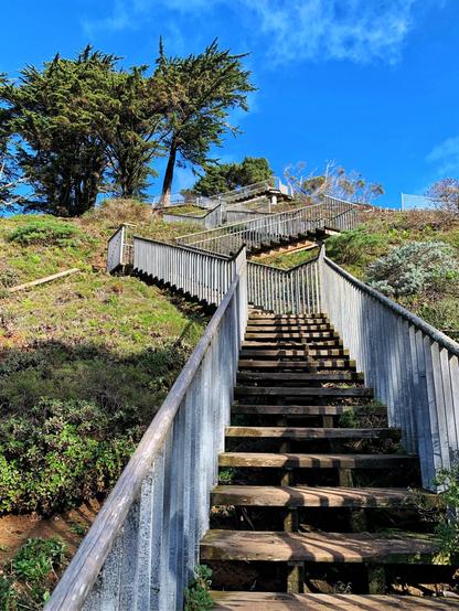 A staircase with metal rails and wooden steps zigzagging up a green hill with a blue sky with slight wisps of clouds and trees in the upper left-hand side of the picture.
