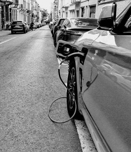 Photo noir et blanc. Vue d'une rue en diagonale.
En haut, à gauche, bâtiments et voitures stationnées.
Puis la rue.
Sur la droite des voitures stationnées.
En bas, à droite, gros plan sur la roue et le cordon de recharge électrique qui déborde des tracés sur la route.