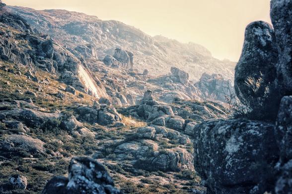A close-up shot of a rocky mountain slope under a hazy sky. The foreground is dominated by large, weathered grey and brown boulders, leading up to a rugged, steep incline. The soft, hazy light from the setting or rising sun washes over the scene, creating a sense of serenity and grandeur.