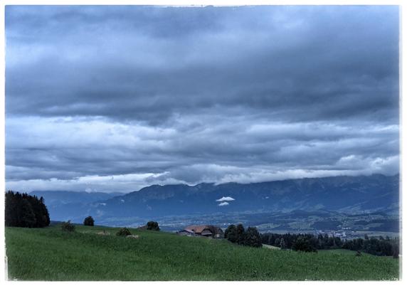A gras field leads into a Valley, another Hill at the horizon. Grey clouds in the sky.
