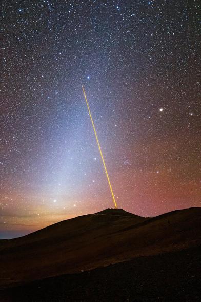 A night landscape in a desert. A bright, thin yellow line extends from the top a black mountain into the starry sky. The yellow laser meets with a blurry streak of white that extends from the horizon, forming an upside-down V. The sky just above the mountain is orange and pinkish, with thousands of bright white stars speckling the night sky.