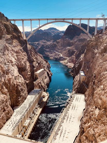 A panoramic view of the Hoover Dam and the Colorado River, featuring a large bridge arching over a dramatic canyon landscape, with hydroelectric power facilities visible along the riverbanks.