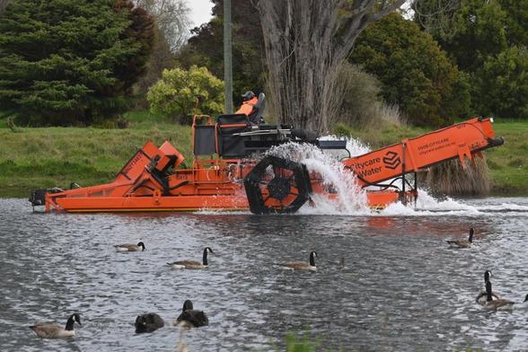 A photo of a man sitting on the top of an enormous orange metal paddle boat churning its way up a river. I photographed this today on the Avon-Ōtākaro river in Christchurch, NZ. It doesn't just look wicked cool but it also cleans up the river's water weeds.

There's a group of Canada geese on the river.