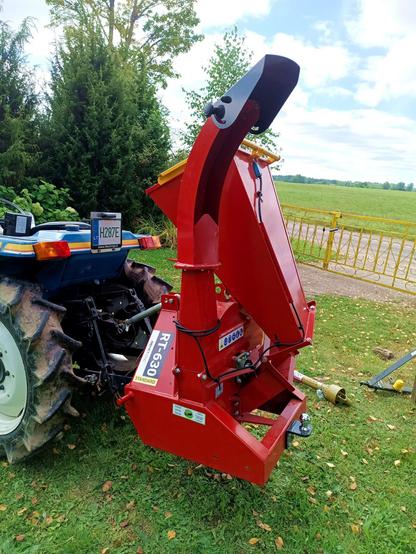 A red wood chipper that is attached to the three point linkage of a small blue Iseki tractor. It says RT-630 on it and the big steel funnel is folded back for transport. It has an exit chute that can be rotated with a black steel deflector for adjusting the height at which it spits out chips. It also has a very handy trailer hitch on the back of it. A yellow PTO shaft lies on the ground and part of a removed drawbar.