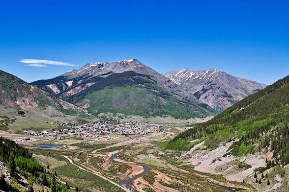 View from above a wide alpine basin. In the foreground a wide flood plain and meandering river; in middle distance the grid of a small town behind which are large mountains with forested slopes and bare, rocky summits. Blue sky.
©BosqueBill.com