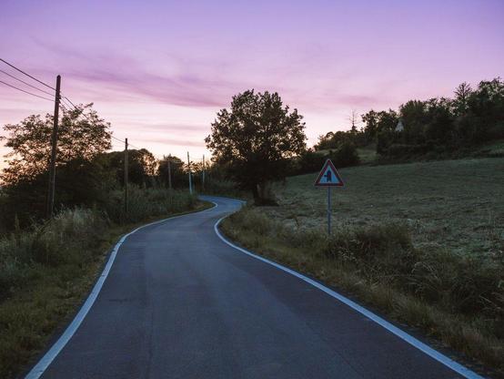A rural road in the foreground winds up a hill with fields and trees on the sides. A street sign is on the right, and a line of telephone poles is on the left, parallel to the road. The sky is pink, blue, and violet.