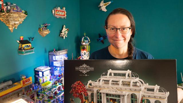 Eine Frau hält eine Schachtel eines Lego Sets in der Hand und lächelt in die Kamera. Im Hintergrund sieht man mehrere Gebäude aus Klemmbausteinen.