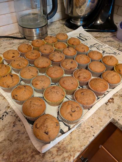A bunch of banana chocolate chip muffins sitting on a black and white tea towel on a granite countertop. There's a black KitchenAid mixer in the background.