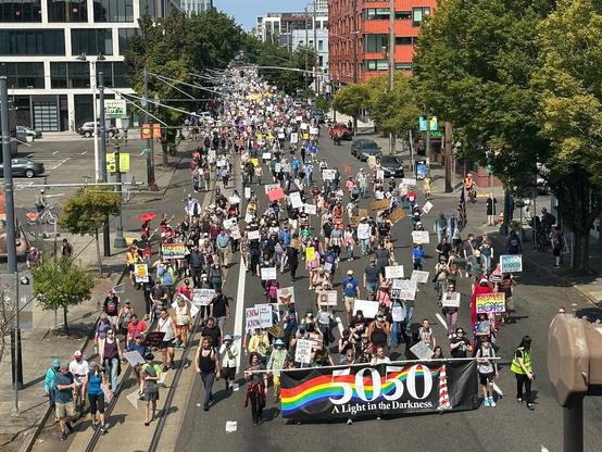 A crowd of people marching down the street in Portland, Oregon. A banner at the front of the march says, "50501: A light in the darkness." The final 1 in 50501 is shaped like a lighthouse.