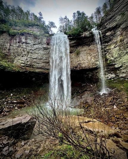 A scenic view of two waterfalls cascading down rocky cliffs, surrounded by greenery and sparse vegetation. The water flows into a small pool at the base, amidst stones and pebbles.