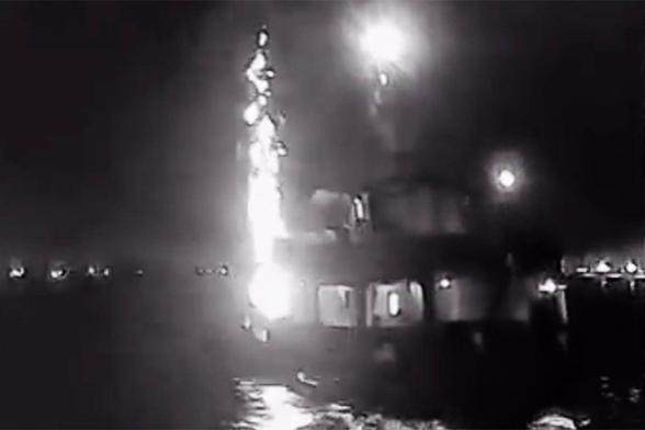 The image of the Israeli terrorist attack on the ship  named 'Family' of the Global Sumud Flotilla depicts a nighttime scene with a large vessel, likely a ship, on water. The vessel is illuminated by bright lights, with a prominent light source on its upper deck. A bright, vertical plume of light and smoke is erupting from the ship, suggesting a missile launch or explosion. The plume is highly luminous, with multiple bright spots indicating the intensity of the event. The surrounding water reflects the light, creating a shimmering effect. The background is dark, with faint lights visible in the distance, possibly indicating other vessels or structures. The overall scene is captured in black and white, emphasizing the contrast between the bright plume and the dark surroundings.