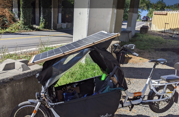 Cargo bike with a solar panel laid on top of the rain canopy and a 200Wh solar inverter battery pack hanging inside the cargo basket, shipping container and a bike repair stand in the background, with a sign for the tool library.  Sunny and blue sky.