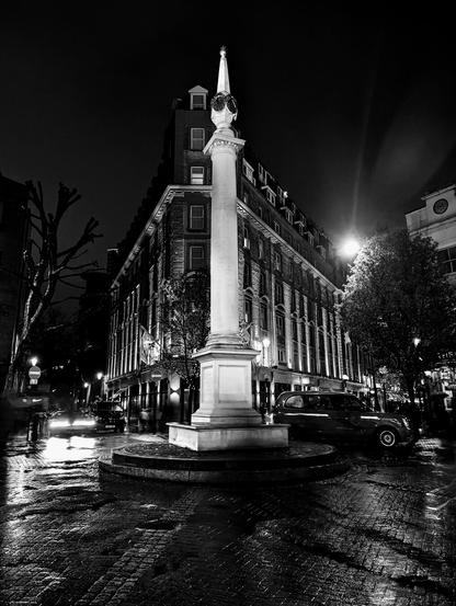 A striking black and white photo of a column monument in a circular cobbled square at night. The monument, the Seven Dials Sundial Pillar, is illuminated from below, highlighting its ornate base and column topped with six sundial faces and a small obelisk. A traditional London taxi is parked to the right. A multi-storey Georgian building with many windows and streetlights is visible behind the monument. The wet cobblestones reflect the lights, creating a shimmering effect. The atmosphere is quiet, with a hint of motion blur from a person on the left and a vehicle in the distance.