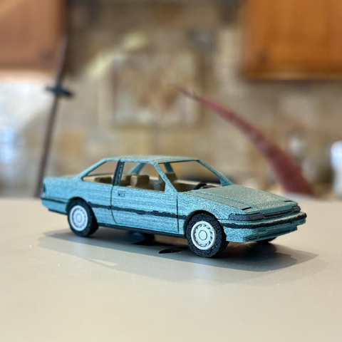 A metallic turquoise 3D printed 1:64-scale 1989 Honda Accord coupe sits atop a closed silver laptop computer, seen from a front-3/4 angle.