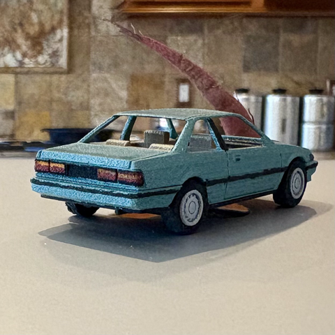 A metallic turquoise 3D printed 1:64-scale 1989 Honda Accord coupe sits atop a closed silver laptop computer, seen from a rear-3/4 angle.