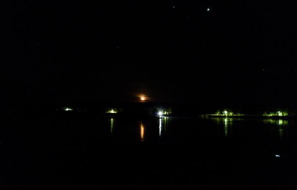 Looking over the ocean, the fingernail moon is orange and reflecting off the surface of the ocean. 5 white human lights ruin the darkness, but there are still stars visible above. Time is 0339h AST
