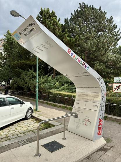 A bicycle station featuring a stylized structure with information panels and a bike parking rack. It is situated near a paved path, with greenery in the background and a car parked nearby. The structure includes graffiti and signage related to local bike services.
