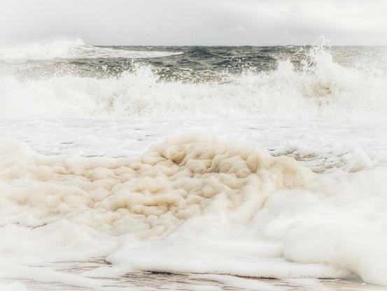 A close-up, low-angle shot of a large pile of thick, yellowish-white sea foam on a sandy beach. In the background, large, dark grey waves crash towards the shore under a cloudy, white sky. The image has a slightly desaturated, moody tone.