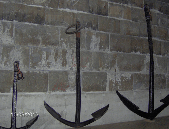 Photo of three anchors, made of dark metal, left to right is smaller to bigger, placed against a large brick wall.