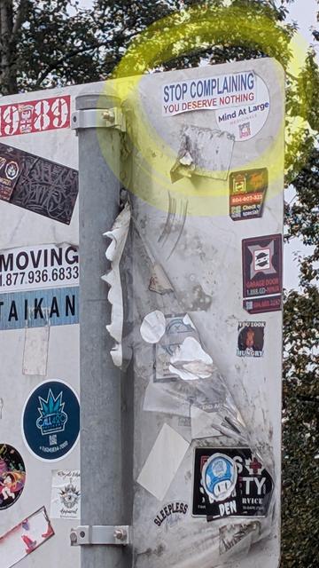 The back of a traffic sign in a left hand turn lane in Burnaby BC has many anarchic stickers on it for waiting drivers to read.