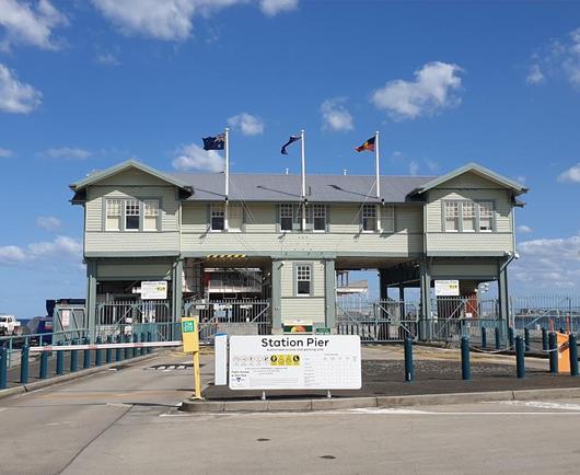 A weatherboard single story building on stilts several metres high. The building is built across a pier. Three flags fly from flagpoles on the building.A sign in front says Station Pier.