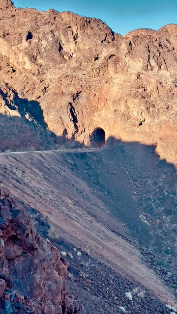 A rugged mountain landscape featuring rocky terrain and a shadowed tunnel entrance. A winding dirt road is visible along the slope. The scene is illuminated by bright sunlight against a clear blue sky.