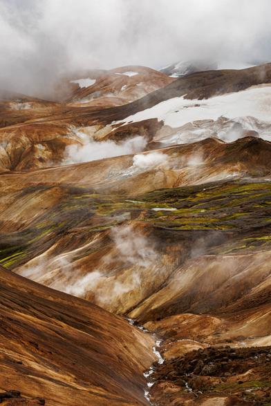 A vertical shot of orange, white, and green geothermal hills with steam pockets and snow here and there.