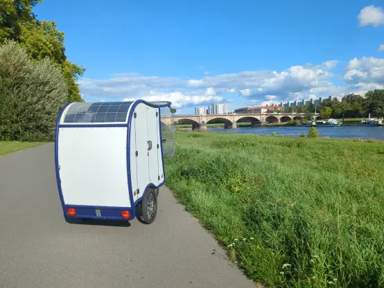 Das Heck der Lastenrad-Kabine wird von der Sonne angestrahlt. Auf dem abgerundeten Dach sind zwei Solarpanele installiert. Es steht auf dem Elbradweg, der Blick geht elbaufwärts, mit der Sonne.