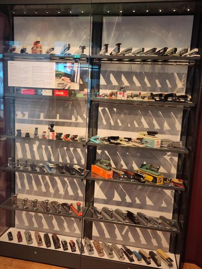 Shelves of staplers of different shapes, sizes and colours displayed in a glass cabinet in a museum. There's strong light from the top left that casts shadows of the staplers and shelving on the white wall behind. Also to the top left is a description of the exhibit and a picture of the collector that's partly obscured by reflections.