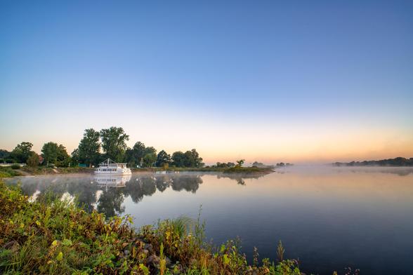 Dieses Bild zeigt einen friedlichen Morgen am Ufer eines breiten Flusses. Eine leichte Nebelschicht liegt über dem Wasser und den fernen Ufern, was eine ruhige und leicht mystische Atmosphäre schafft.
Im Zentrum des Bildes liegt ein weißes Ausflugsschiff, das auf dem ruhigen Wasser ankert. Seine Spiegelung ist deutlich auf der glatten Oberfläche zu sehen. Der Himmel ist klar und geht von einem sanften Hellblau im oberen Bereich in einen warmen, orange-gelben Ton über, der den Sonnenaufgang andeutet.
Am linken Bildrand steht eine Gruppe von Bäumen mit dichtem Laubwerk, die ebenfalls im Wasser reflektiert wird. Im Vordergrund sind Pflanzen und hohes Gras zu sehen, das teilweise mit Morgentau bedeckt ist.
Die Komposition des Bildes mit dem breiten Fluss, dem Schiff, den Bäumen und dem weiten Himmel strahlt eine tiefe Ruhe und Stille aus. Die sanften Farben und die spiegelglatte Wasseroberfläche machen die Szene besonders harmonisch.